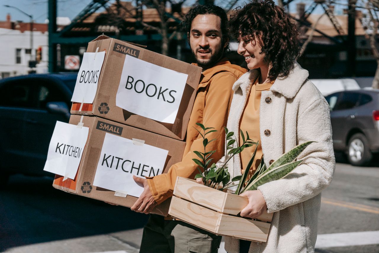 Joyful couple carrying boxes and plants while relocating to a new neighbourhood on a sunny day.