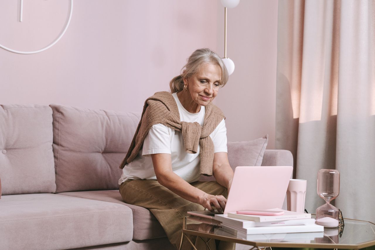 Elderly woman with grey hair smiling while working on a laptop at home, enjoying remote work.