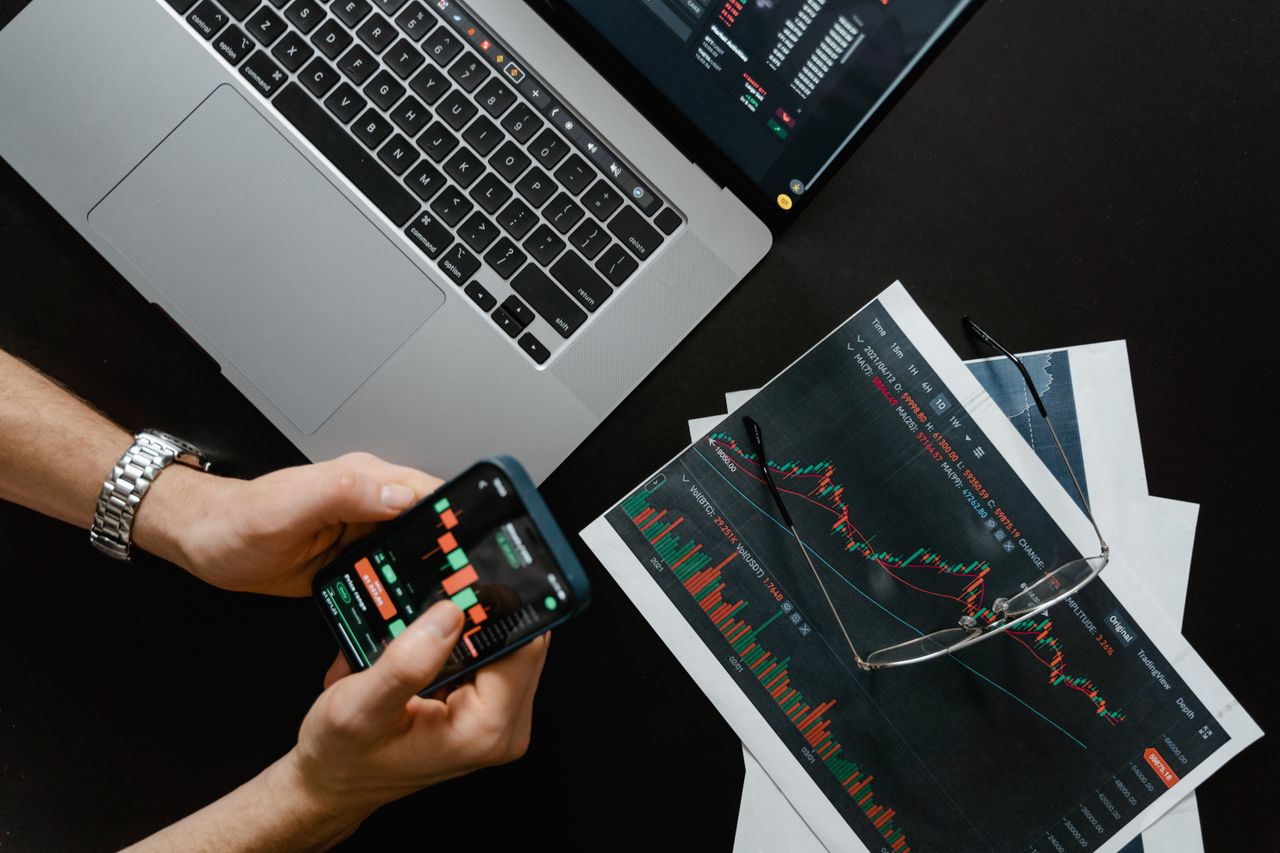 An overhead desk with a laptop, phone and a candlestick trading chart.